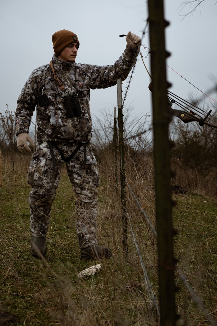 A person in camouflage and tactical gear adjusting a fence in an outdoor field.